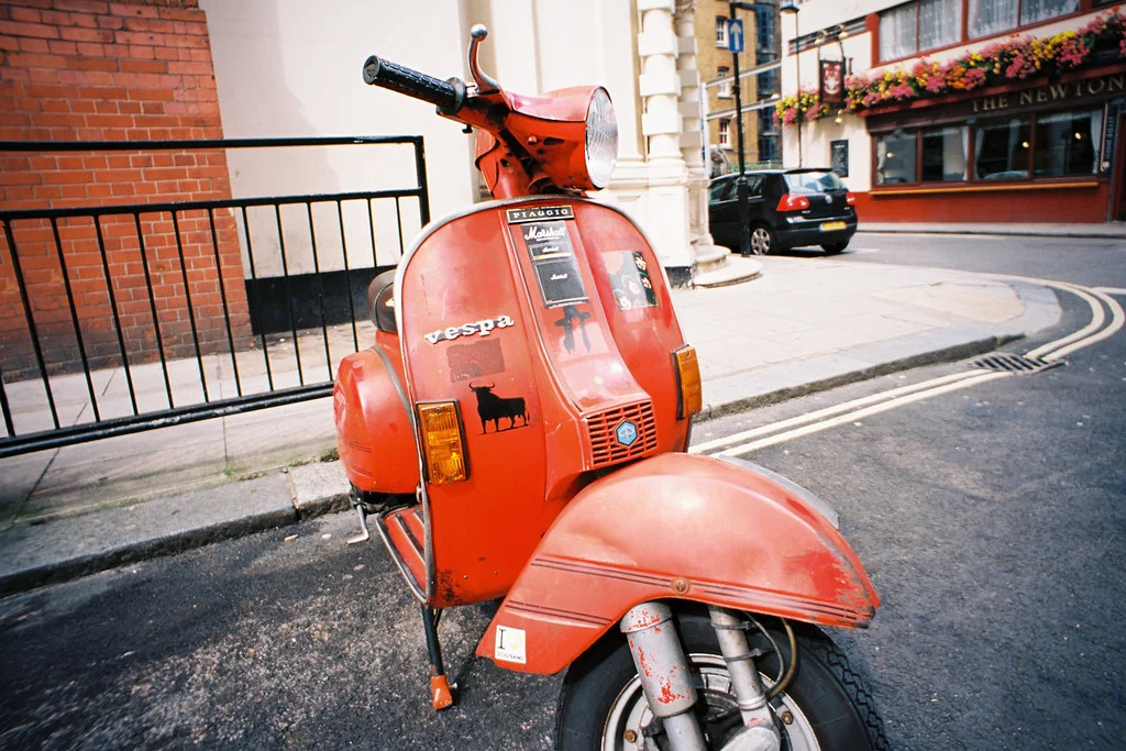 Vespa Scooter: A step-through scooter with a rounded front shield, narrow wheels, curved body, and partly enclosed mechanics.